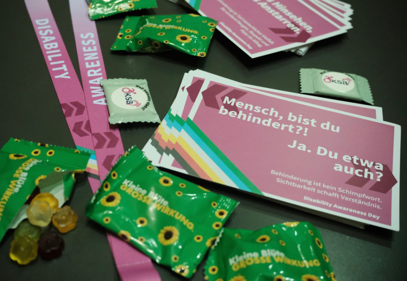 Giveaways: Flyer, Schlüsselband, Buttons und verpackte Süßigkeiten liegen auf dem Stand-Tisch von KSB/VSUZH mit Informationsmaterial zum Disability Awareness Day.